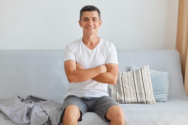 Indoor shot of smiling positive man wearing casual style white t shirt sitting on sofa looking at camera keeping hand folded expressing positive emotions