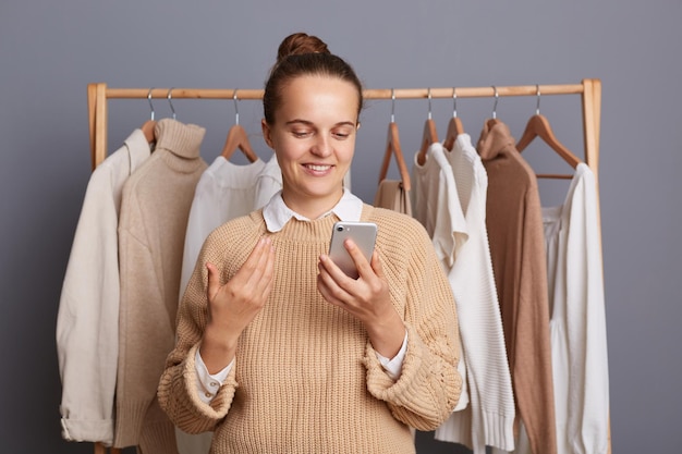 Снимок в помещении улыбающейся позитивной женщины, стоящей возле вешалки с одеждой в модном магазине и держащей смартфон в руках, болтающей с друзьями, хвастающейся своими покупками