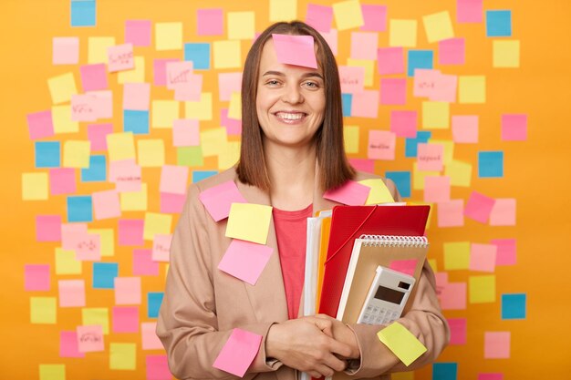 Indoor shot of smiling satisfied attractive Caucasian woman wearing beige jacket posing against yellow wall with colorful memo cards holding paper folder looking at camera being in university