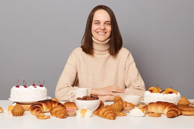 Tiro al coperto di una donna soddisfatta sorridente che indossa un maglione con i capelli castani seduti a tavola con una deliziosa pasticceria fatta in casa guardando la fotocamera essendo affamata isolata su sfondo grigio