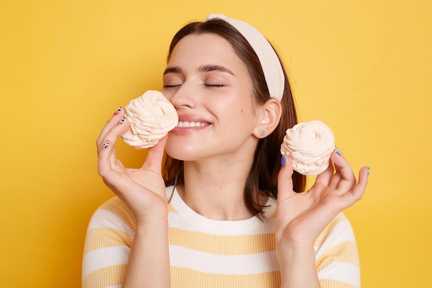 Indoor shot of smiling satisfied woman wearing striped shirt standing with closed eyes and smelling cake enjoying delicious dessert posing isolated over yellow background