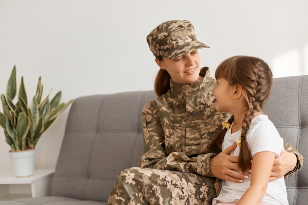 Indoor shot of smiling soldier female sitting and looking at\
her kid with smile being happy to return home from army woman\
wearing camouflage uniform and cap posing with her daughter