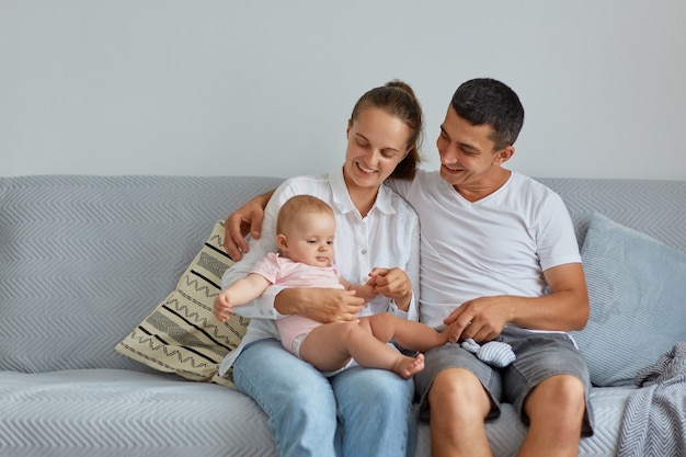 Indoor shot of smiling wife and husband wearing casual clothing sitting on sofa with charming infant daughter in light room, spending happy time together.