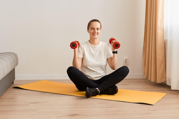 Indoor shot of smiling woman exercising with dumbbells at home sporty beautiful young girl training at home to stay fit female having work out in living room