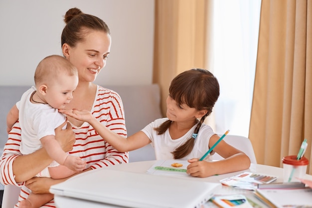 Indoor shot of smiling woman holding infant baby and helping her elder daughter with homework sitting at table and writing posing in light living room