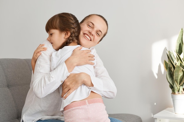 Indoor shot of smiling woman hugging her daughter, family expressing love and happiness, being glad to spend time together at weekend, motherhood and childhood.