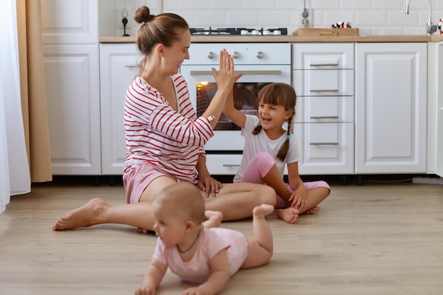 Indoor shot of smiling woman wearing striped casual style shirt sitting on floor in kitchen with her children cute little baby crawling mother giving five to her daughter