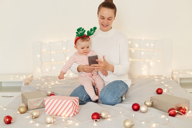 Indoor shot of smiling woman wearing white sweater and jeans posing with infant daughter on bed with Christmas decoration, holding cell phone in hands, writing messages.