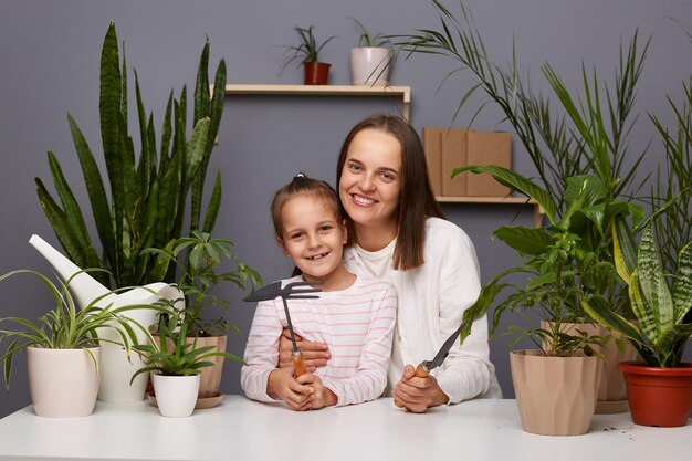 Indoor shot of smiling woman with dark hair posing with cute little daughter in room with greenery sitting at table taking care of flowers looking at camera