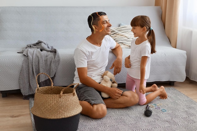 Indoor shot of smiling young adult father spending time with his daughter while sitting on floor near sofa in living room, kid making up lips with lipstick for daddy and admires her result.