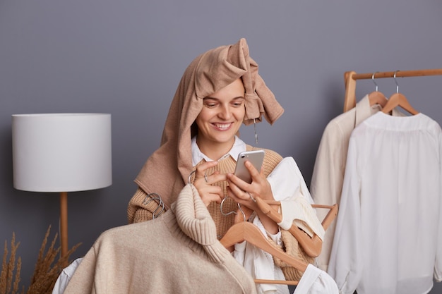 Снимок в помещении улыбающейся молодой взрослой женщины, держащей вешалки с одеждой и мобильным телефоном, позирующей в модном магазине или выставочном зале, печатающей сообщения на своем мобильном телефоне, продающей одежду