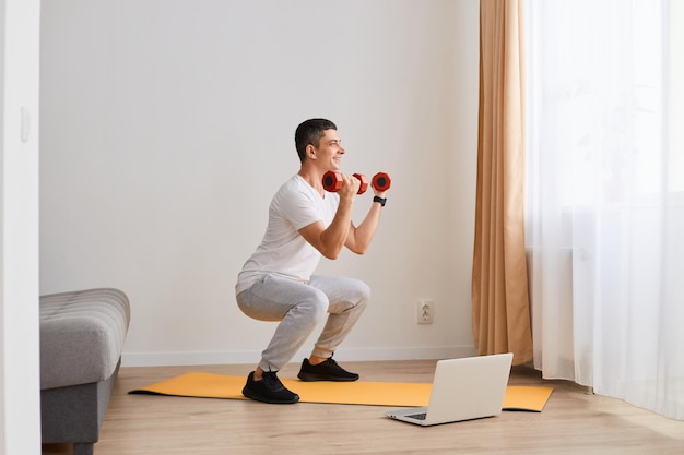 Indoor shot of sporty athletic man wearing sportswear doing sport exercises at home squatting doing situp at home having concentrated look holding dumbbells in hands