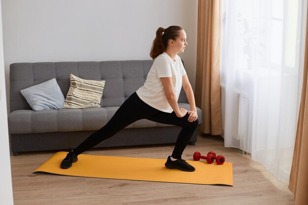 Indoor shot of sporty woman with ponytail wearing black leggins and white t shirt doing sport exercises on yoga mat, warning up her legs, practicing yoga.