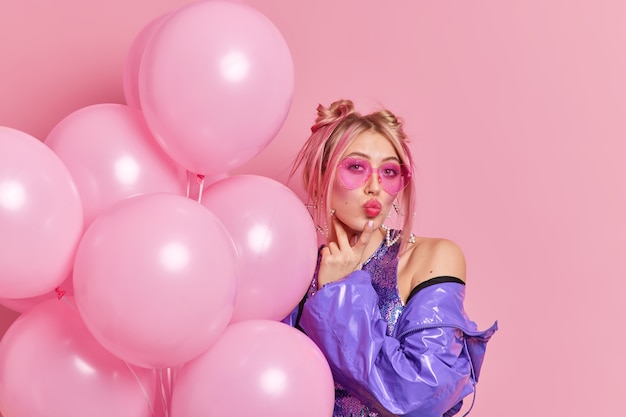 indoor shot of stylish european woman keeps lips rounded wears bright makeup trendy heart shaped sunglasses purple jacket poses with inflated pink balloons comes on birthday party. celebration concept