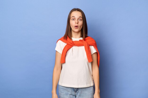 Indoor shot of surprised Caucasian girl wearing white Tshirt and orange sweater tied over shoulders standing against blue wall standing looking at camera with big eyes sees something astonished