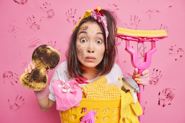 Indoor shot of surprised ethnic housewife holds dirty sponge and mop busy doing housework stands near basket of laundry poses against pink wall