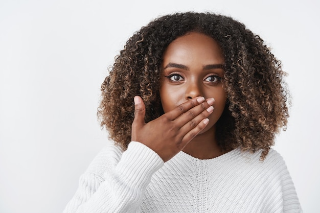 Indoor shot of surprised and stunned woman gasping from shock cover mouth speechless and amazed looking worried at front reacting to shocking unexpected situation over white wall