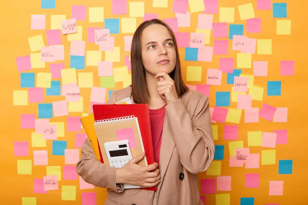 Indoor shot of thoughtful woman student with brown hair wearing beige jacket standing with paper folder holding her chin thinking about her studying posing isolated over yellow background