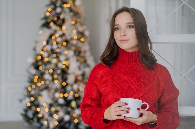 Indoor shot of thoughtful young female in warm winter clothes enjoys hot coffee or cappuccino looks pensively aside stands near decorated Christmas tree has red manicure Coziness concept