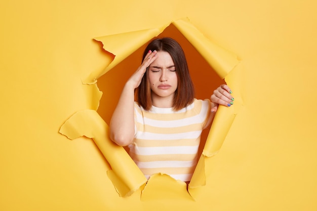 Indoor shot of tired exhausted young adult brunette woman stands in torn paper hole suffering headache touching head looking through breakthrough of yellow background