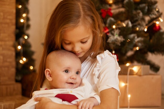 Indoor shot of two sisters posing near Christmas tree at home, elder girl hugging infant baby with love, merry Christmas and happy new year, children in festive room.