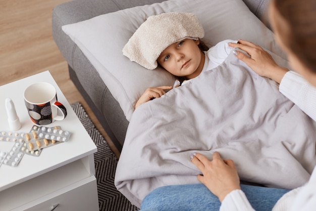 Indoor shot of unhealthy ill female child with wet towel on her forehead lying on sofa under blanket, mother sitting near her little sick girl, taking care and help.