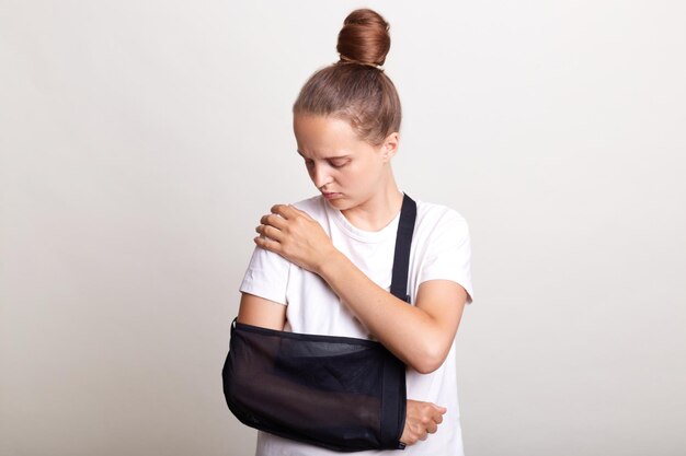 Indoor shot of unhealthy woman wearing casual tshirt standing\
in arm sling and feeling pain in shoulder touching painful part of\
the body car accident posing isolated on light gray background