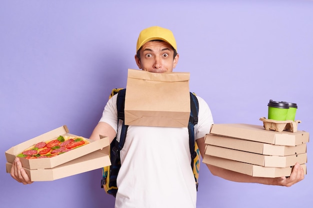 Indoor shot van drukke bezorger in gele pet en tshirt met pizza in kartonnen afhaalmaaltijden koffie en papier pakket in zijn mond heeft veel bestellingen poseren geïsoleerd op violette achtergrond