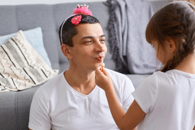 Indoor shot van grappige knappe man met donker haar en roze haarspeld en hoepel met kroon, schattig dochtertje achterstevoren poseren en make-up maken voor papa, pommade gebruiken voor lippen.