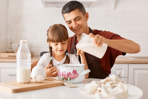 Indoor shot van jonge volwassen donkerharige man met kastanjebruin t-shirt en schort koken in de keuken met dochtertje, melk gieten, deeg voorbereiden voor zelfgemaakt gebak, verrassing voor moeder.