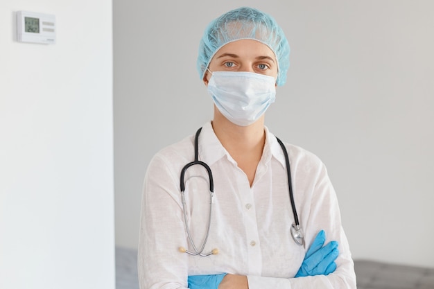 Indoor shot van jonge volwassen mooie vrouwelijke arts met medisch uniform, pet en chirurgisch masker, staande met gekruiste armen en zelfverzekerde gezichtsuitdrukking.