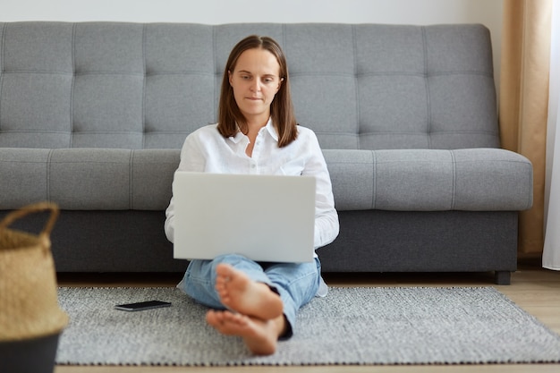 Indoor shot van jonge volwassen vrouwelijke freelancer die online werkt, zittend op de vloer in de buurt van grijze bank, kijkend naar pc-computerscherm, vrouw met wit overhemd en spijkerbroek.