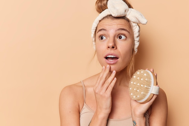 Indoor shot van verraste jonge Europese vrouw houdt droge borstel geniet van dagelijkse ochtendroutine in badkamer draagt hoofdband geïsoleerd over bruine achtergrond met kopieerruimte voor uw promotionele inhoud