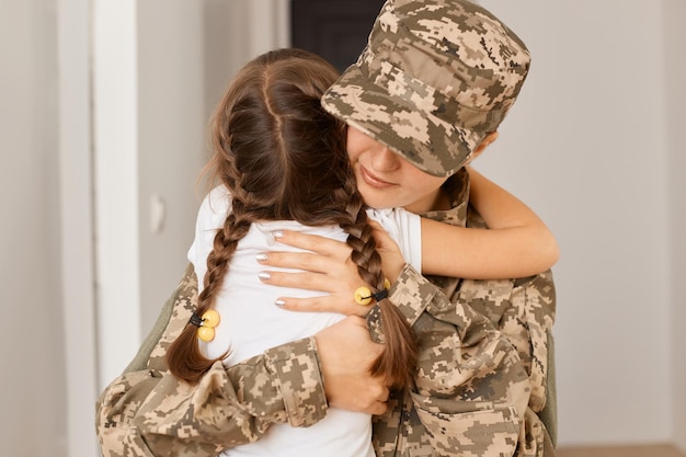 Indoor shot of woman soldier wearing camouflage uniform and cap hugging her little dark haired daughter with braids before going to army or war expressing sadness