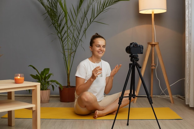 Indoor shot of woman trainer wearing white tshirt shooting\
video lesson on a camera on a tripod while sitting on a yoga mat\
holding glass in hands telling to her followers about water\
balance