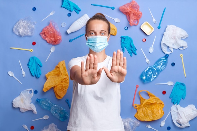 Indoor shot of woman volunteer wearing white t shirt and mask cares about environment cleans from garbage concerned with plastic contamination raises her palms showing stop gesture