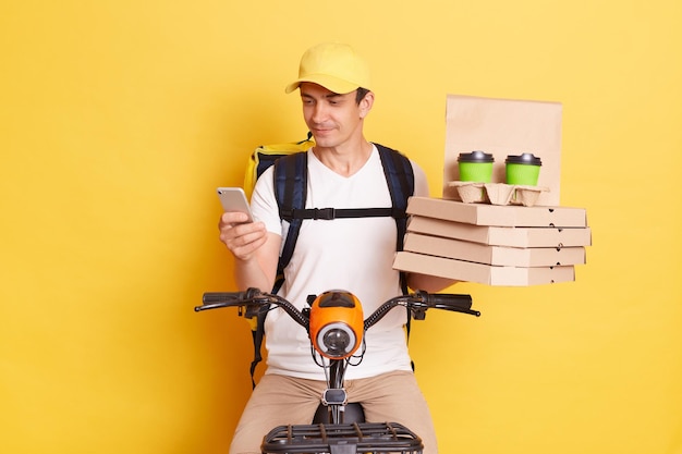 Indoor shot of young adult Caucasian delivery man using smartphone checking address for delivering order wearing cap and thermo bag riding bicycle isolated on yellow background