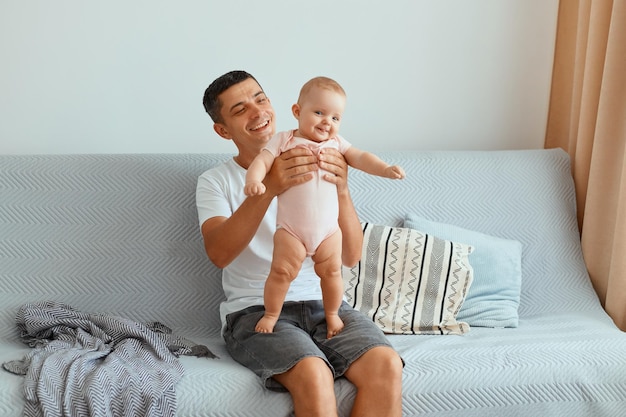 Indoor shot of young adult dark haired man wearing white t shirt and jeans short sitting on cough, happy father holding infant daughter in hands, expressing positive emotions.