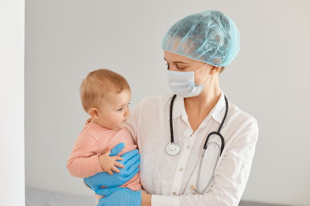 Indoor shot of young adult female pediatrist wearing medical uniform, holding infant baby girl, examining child's health in clinic, being happy the kid benefited from the treatment.