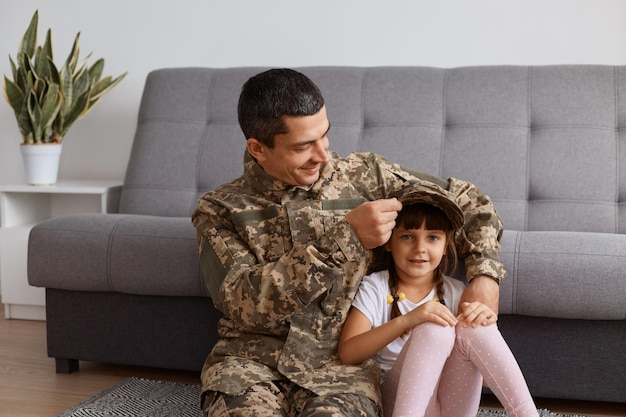 Indoor shot of young adult soldier man wearing camouflage\
uniform returning home after army, spending time with daughter,\
wearing his cap on child\'s head, smiling happily.