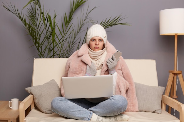 Indoor shot of young adult woman wearing cap gloves scarf and coat sitting in living room with smart phone in hands working online on laptop turning off central heating