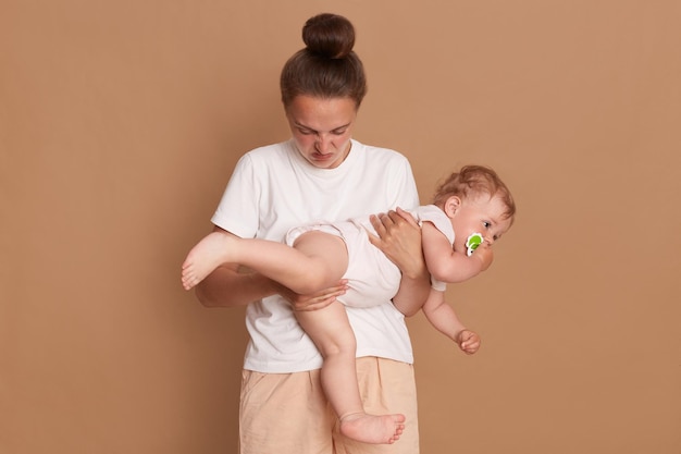 Indoor shot of young adult woman with bun hairstyle wearing white shirt holding baby daughter who crap her pants mother felling bad odor posing isolated over brown background