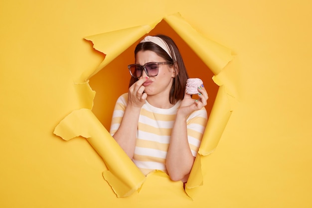 Indoor shot of young dark haired woman wearing hair band and striped t shirt standing in yellow paper hole holding marshmallow and pinching her nose bad odor