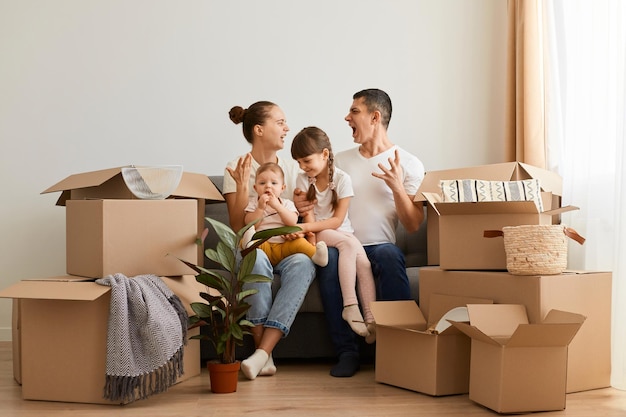 Indoor shot of young family sitting on sofa with children surrounded with carton boxes coupler screaming expressing anger and aggressive having quarrel