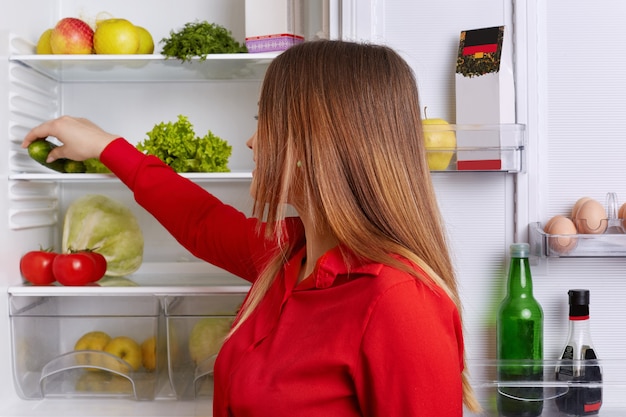 Indoor shot of young female with long straight dark hair, puts vegetables on shelf of refrigerator, eats only healthy food. Woman on kitchen. Housewife going to make vegetable salad.