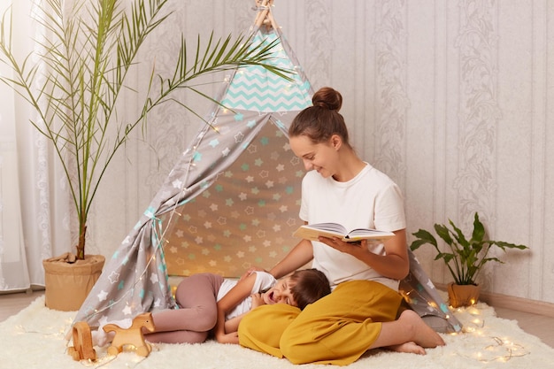 Indoor shot of young mother holding book and talking with\
little daughter female child yawning and falling asleep after\
interesting fairy tale people wearing casual clothing