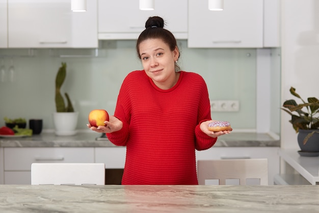 Indoor shot of young pregnant woman with apple and tasty cake in her hands, decides what to eat, attractive female posing in house kitchen.