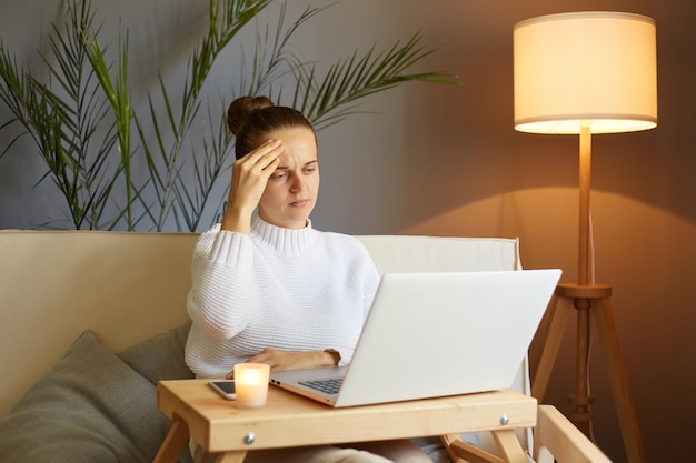 Снимок в помещении молодой женщины, занимающейся исследовательской работой для своего бизнеса, сидя на диване грустной уставшей девушки, просматривающей Интернет в свободное время дома