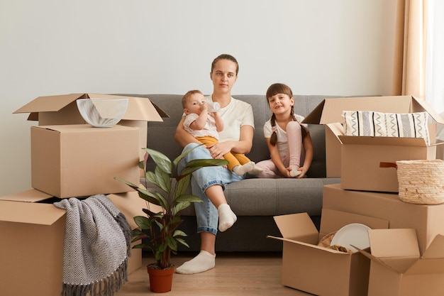 Снимок в помещении: молодая женщина в повседневной одежде сидит на полу со своими детьми и смотрит в камеру, переезжая в новую квартиру, окруженную картонными коробками
