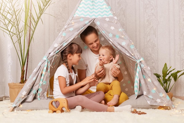 Indoor shot of young woman with two kids sitting in wigwam mother playing with preschool and toddler children clapping hands and smiling happily